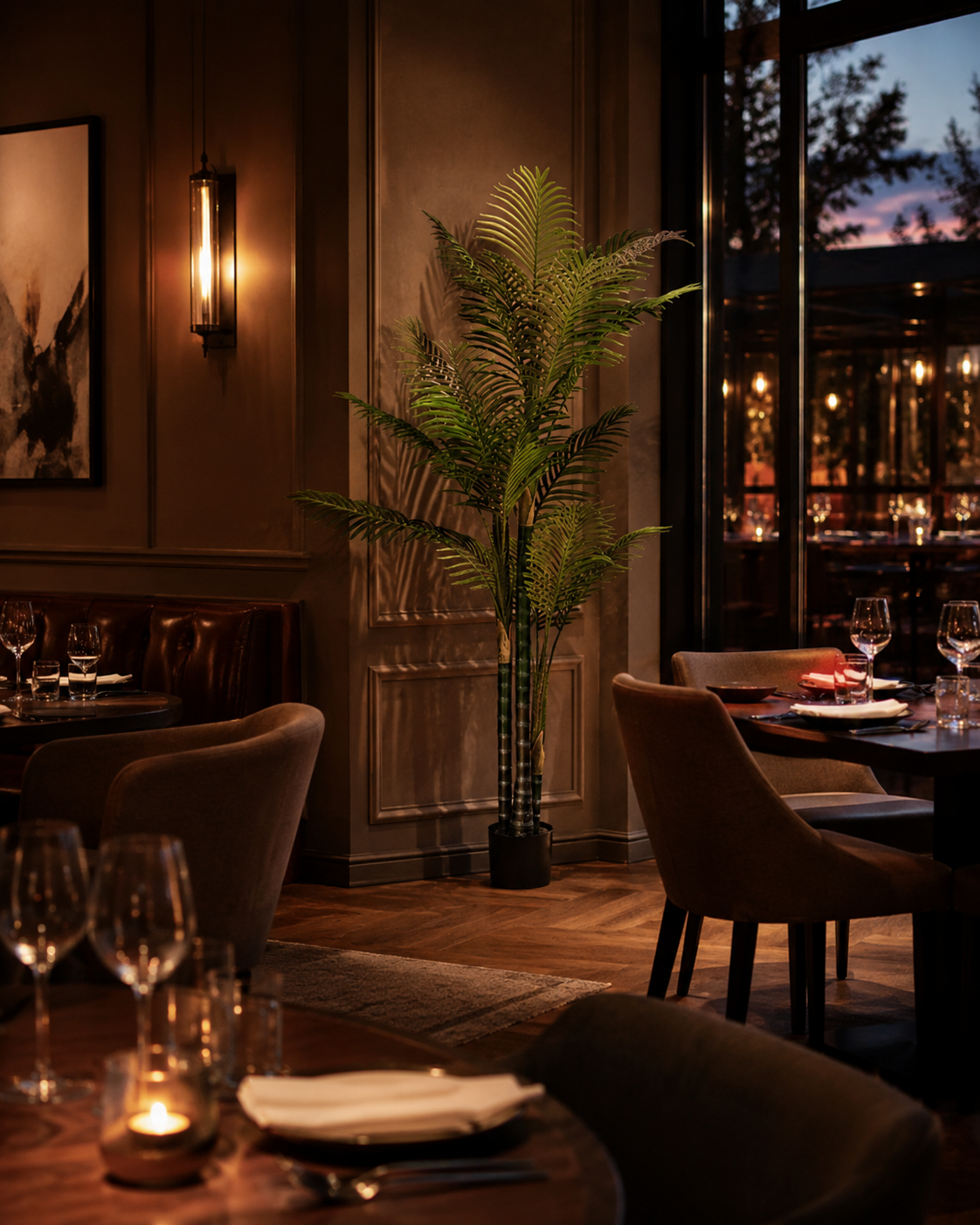 A warmly lit restaurant interior with upholstered dining chairs, tables set with wine glasses and folded napkins, a tall potted palm, and large windows showing an evening outdoor scene.